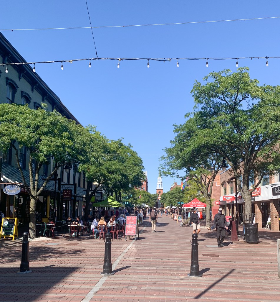 People sitting at outdoor cafes. Trees line the street with a church steeple at the end.