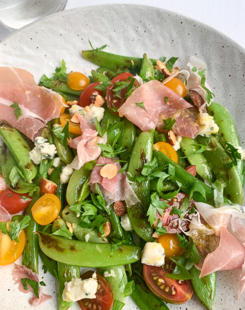 a close-up on the assembled sugar snap salad with ribbons of prosciutto, grape tomatoes, lemon dressing and crumbled blue cheese and chopped almonds.